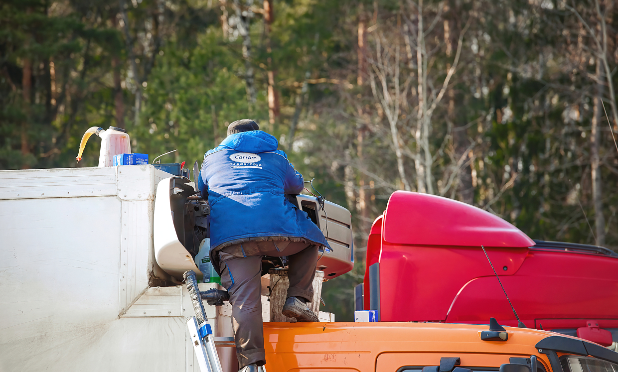 Worker in a blue jacket climbs into the cab of a white utility truck, with an orange boat and red equipment nearby in a forested setting.
