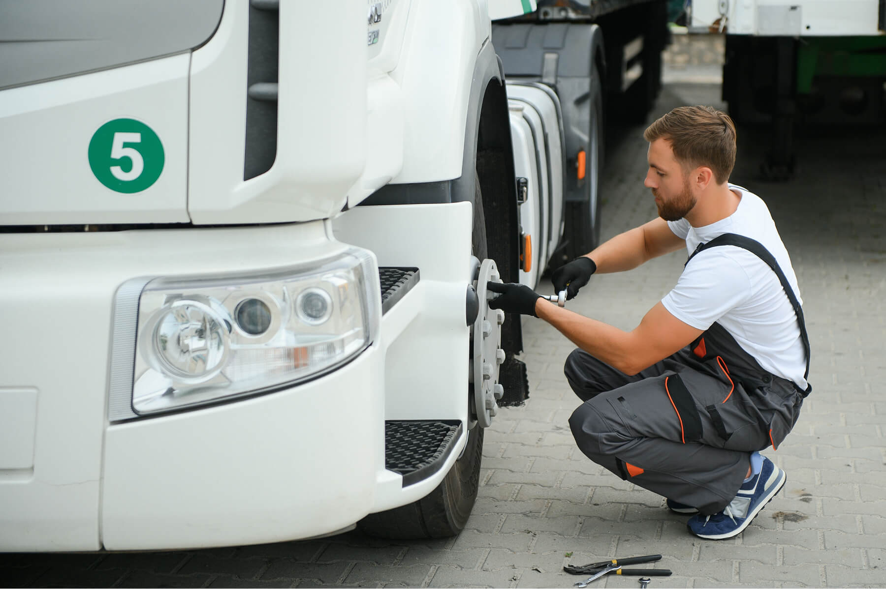 Male mechanic kneels by a white truck, tightening the front wheel with a wrench in his gloved hands.