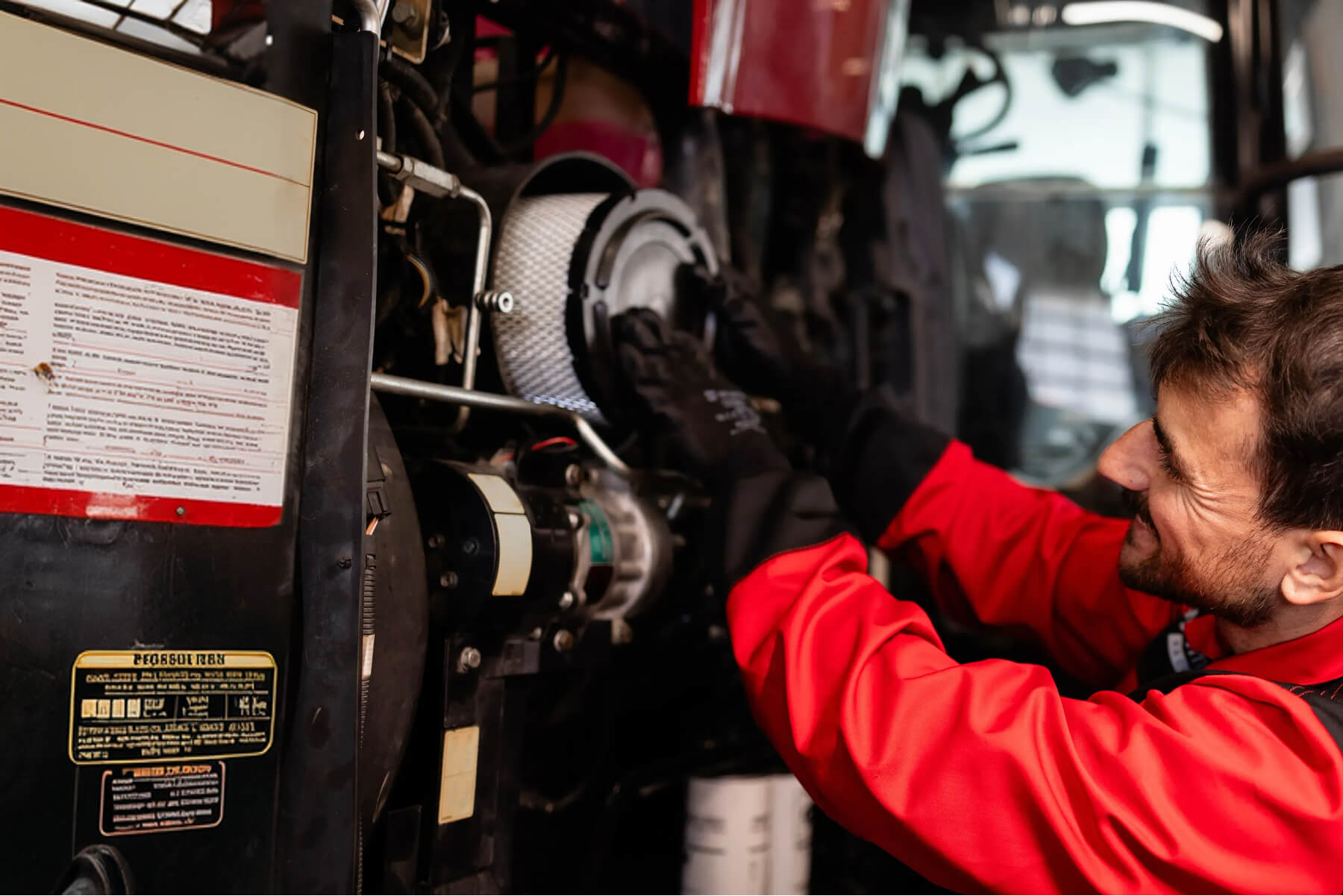 Mechanic in red coveralls inspecting a bus engine, kneeling beside exposed components and labels in a workshop.